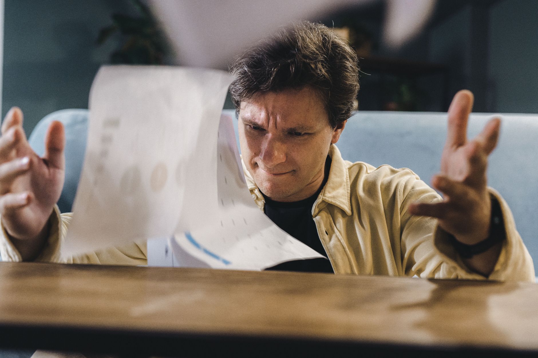 Stressed man throwing documents in frustration at a wooden desk in an indoor setting.