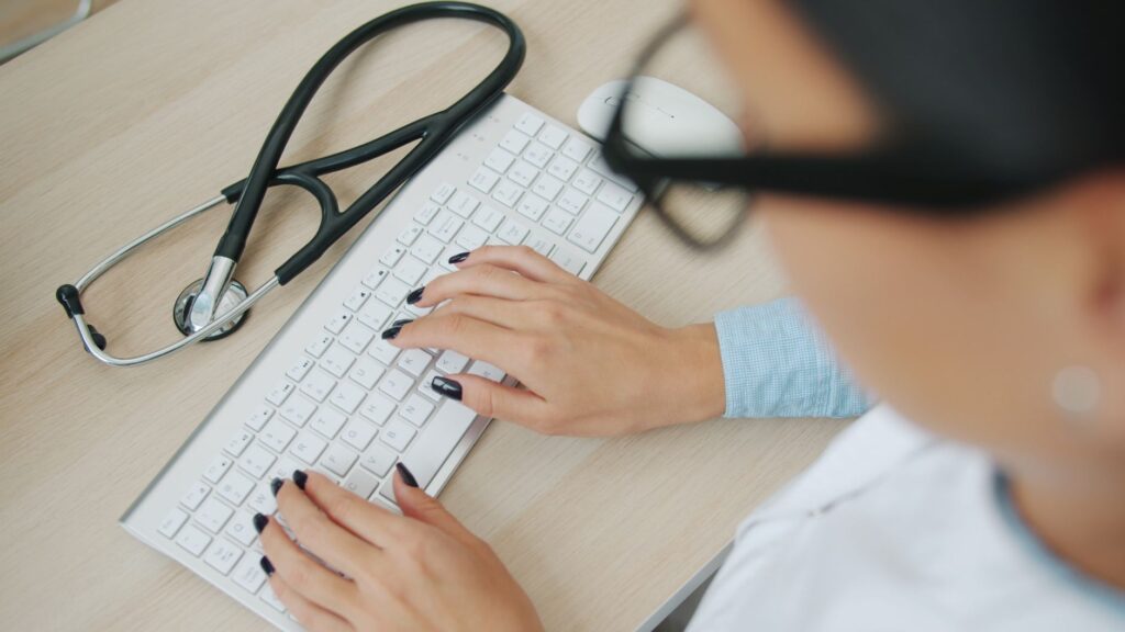 Doctor uses computer keyboard; stethoscope nearby on desk.
